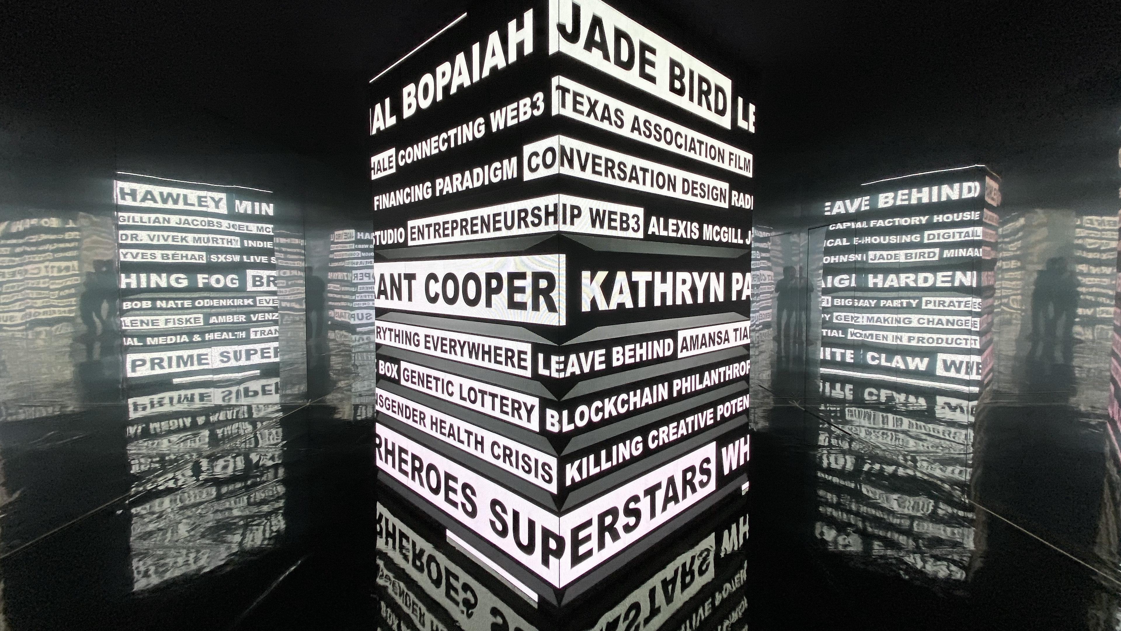 Interactive art installation that consists of an LED Cube that is in the center of a room with mirror walls. The LED cube displays black and white typography listing SXSW 2022 band names.