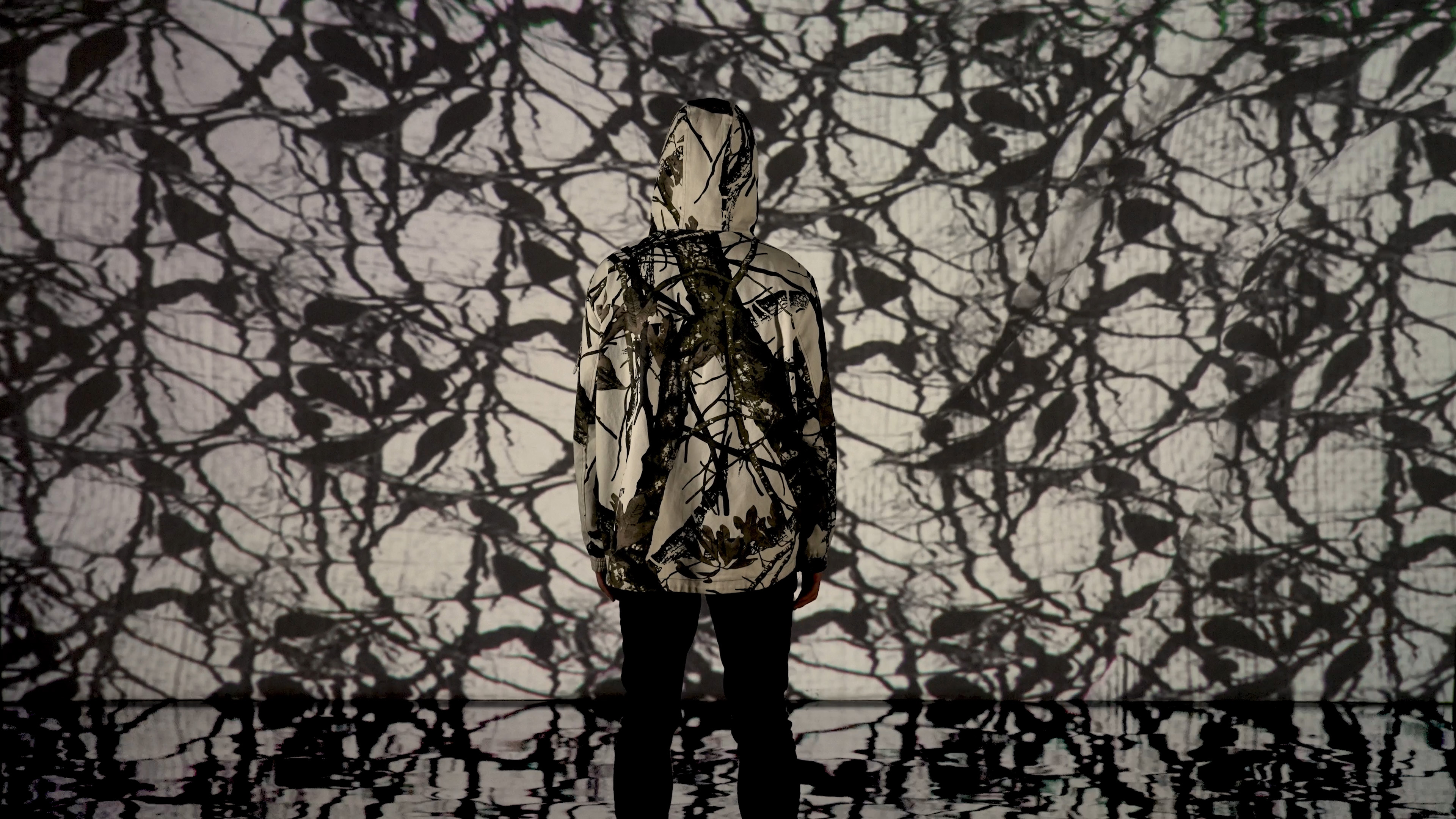 A person wearing a hoodie with a pattern out of tree branches in front of a projection screen that displays a similar pattern out of tree branches