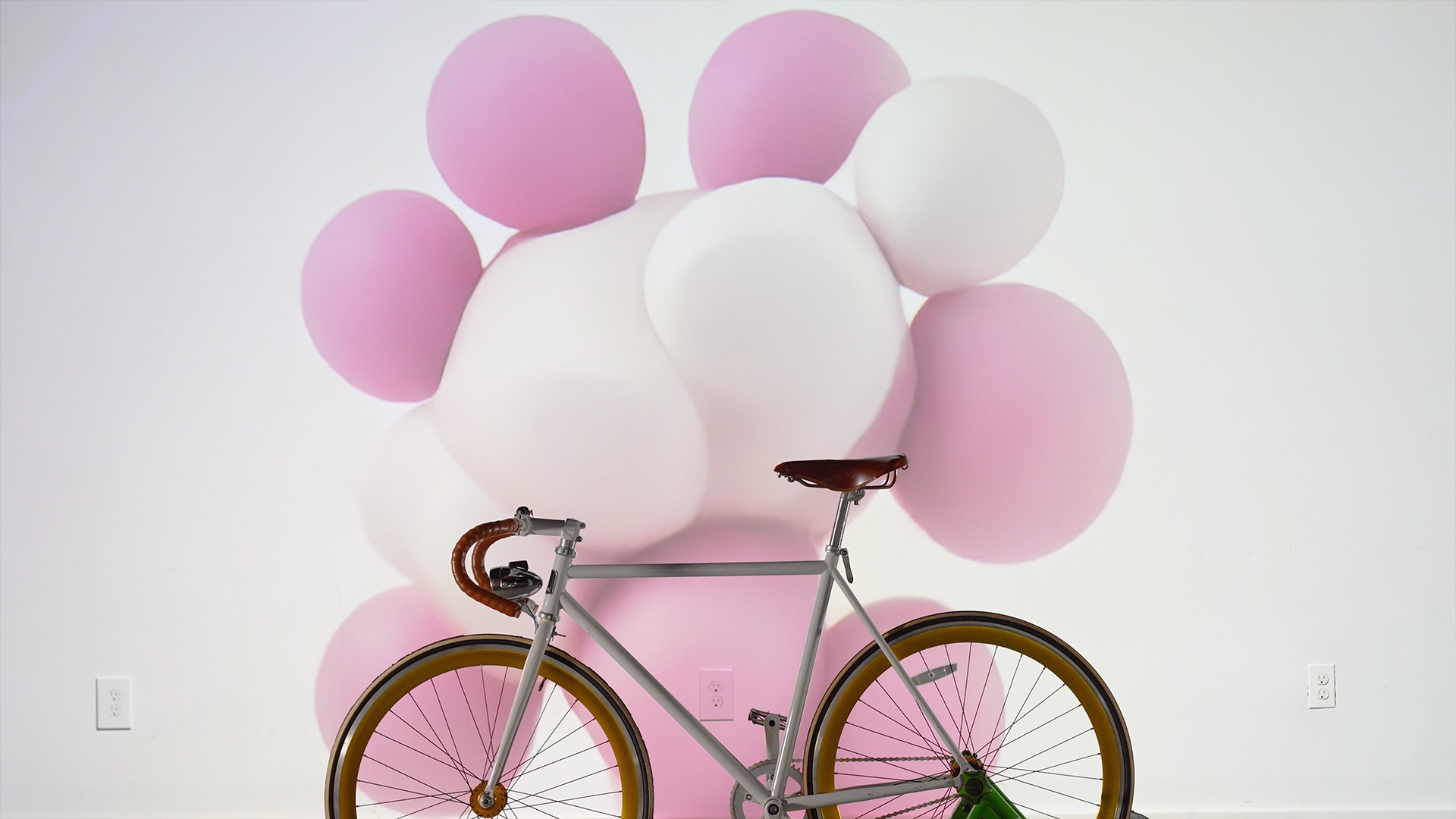 Cycling Dreams Idle state. The image shows a minimalist scene with a white bicycle featuring a brown leather seat and golden-rimmed wheels in front of a white wall displaying ai generated abstract pastel pink and white bubble-like shapes that circle around the bike.