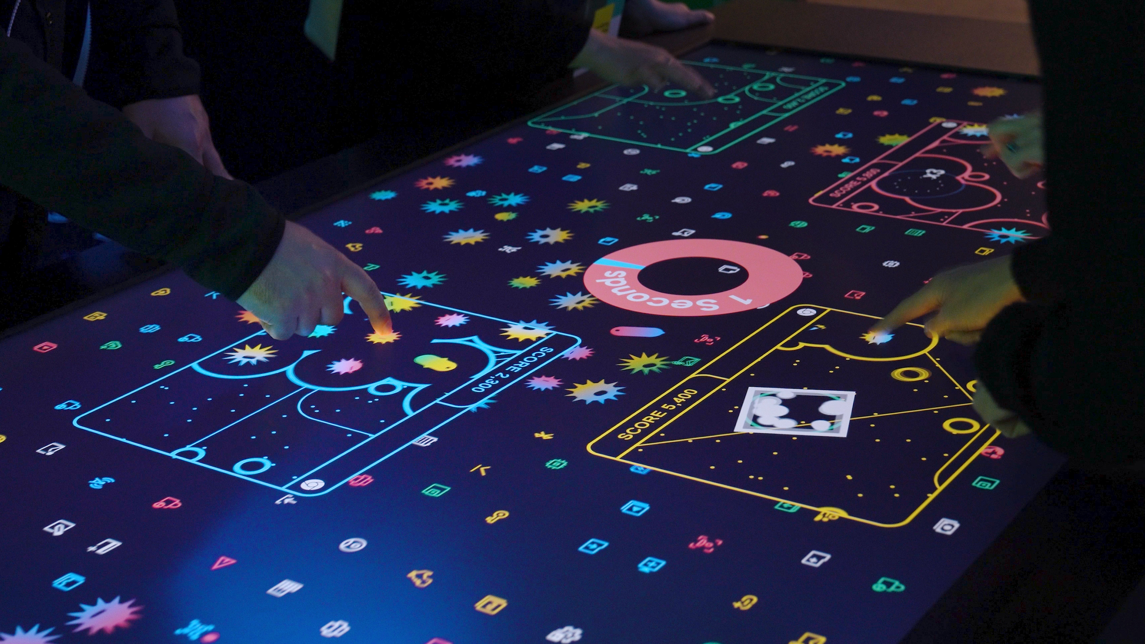 Close up of the touchscreen table display. The Data Defense game experience is in full swing. Four players move around their stylized chrome windows to find and neutralize threats and gain points. All display graphics are very colorful and vibrant.
