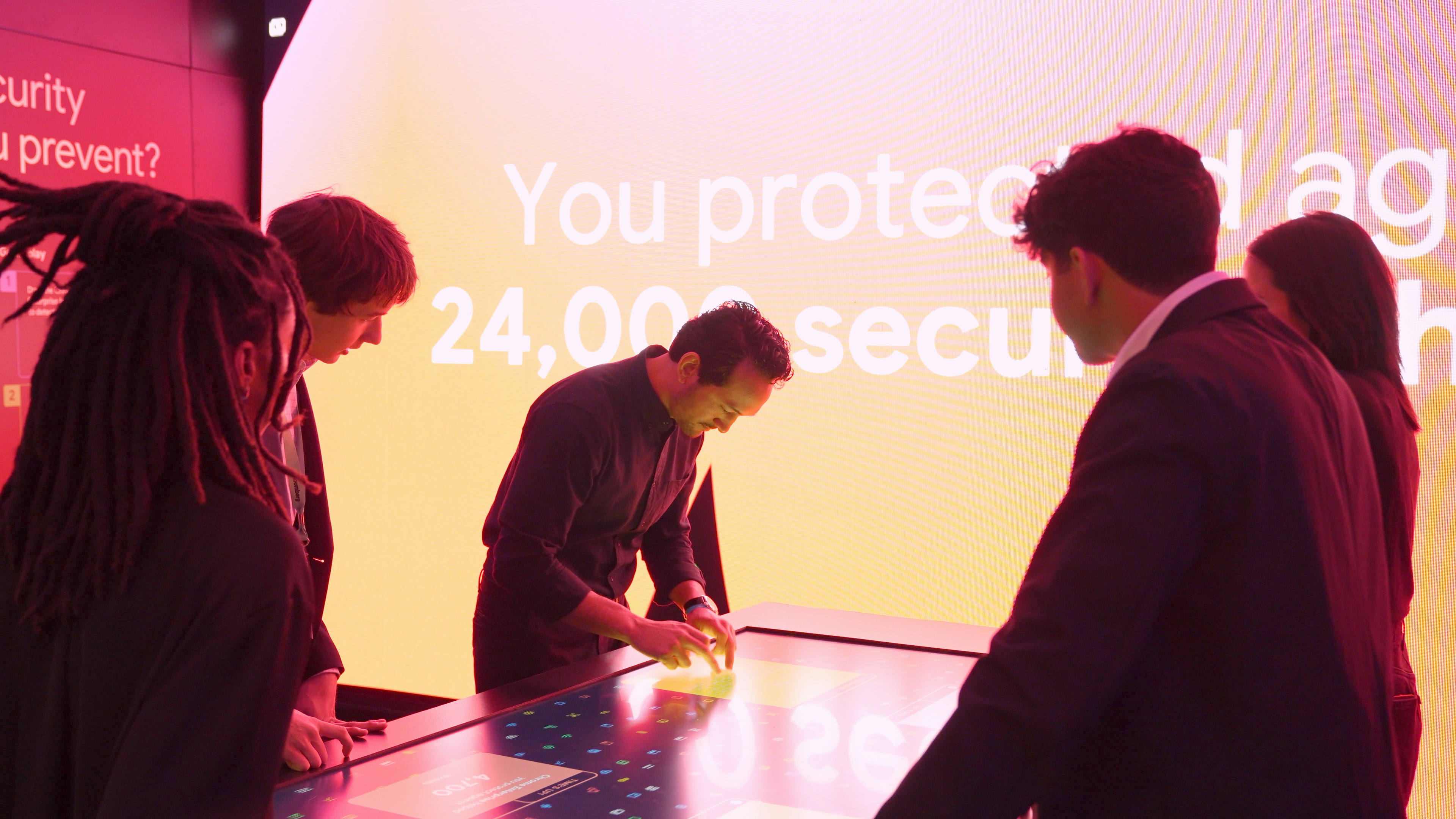 Four Players and a guest stand around the touchscreen table display in front of the big LED screen. The game just finished and one player is entering their team name to submit the score to the leaderboard.