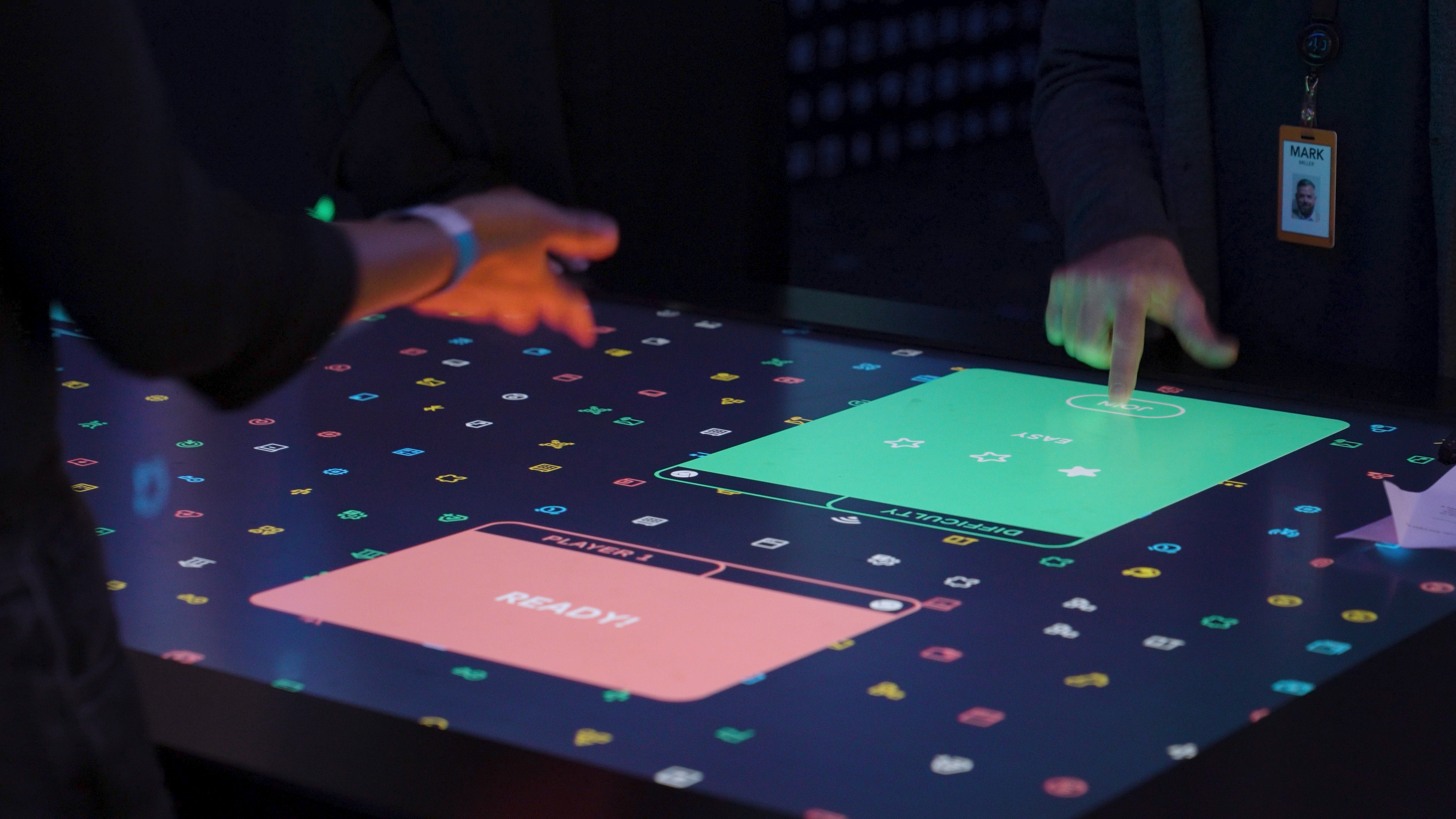 Close up of the touchscreen table display. The playfield of player 1 reads 'Ready'. The hand of a second player touches the join button on the playfield for player 2.
