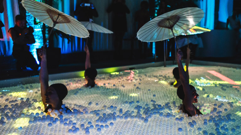 Aqualillies perform a water ballet in a pool covered with ping pong balls and interactive projections