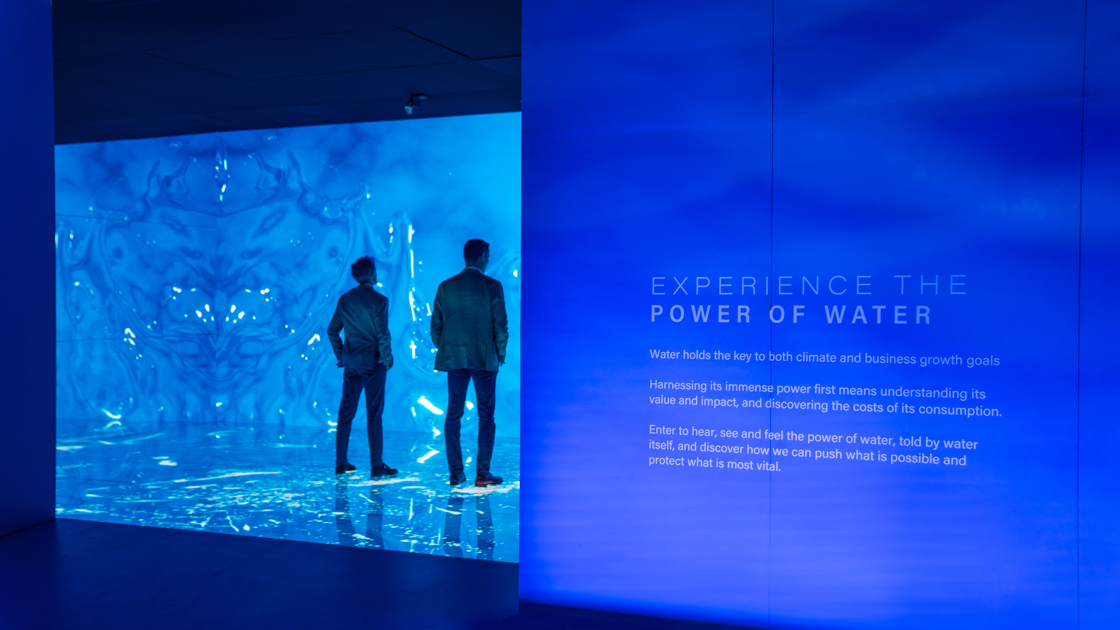 Two people standing inside a room with led walls and floors. Water graphics fill the room. Outside the room is a text description of the installation 'Experience the Power of Water'
