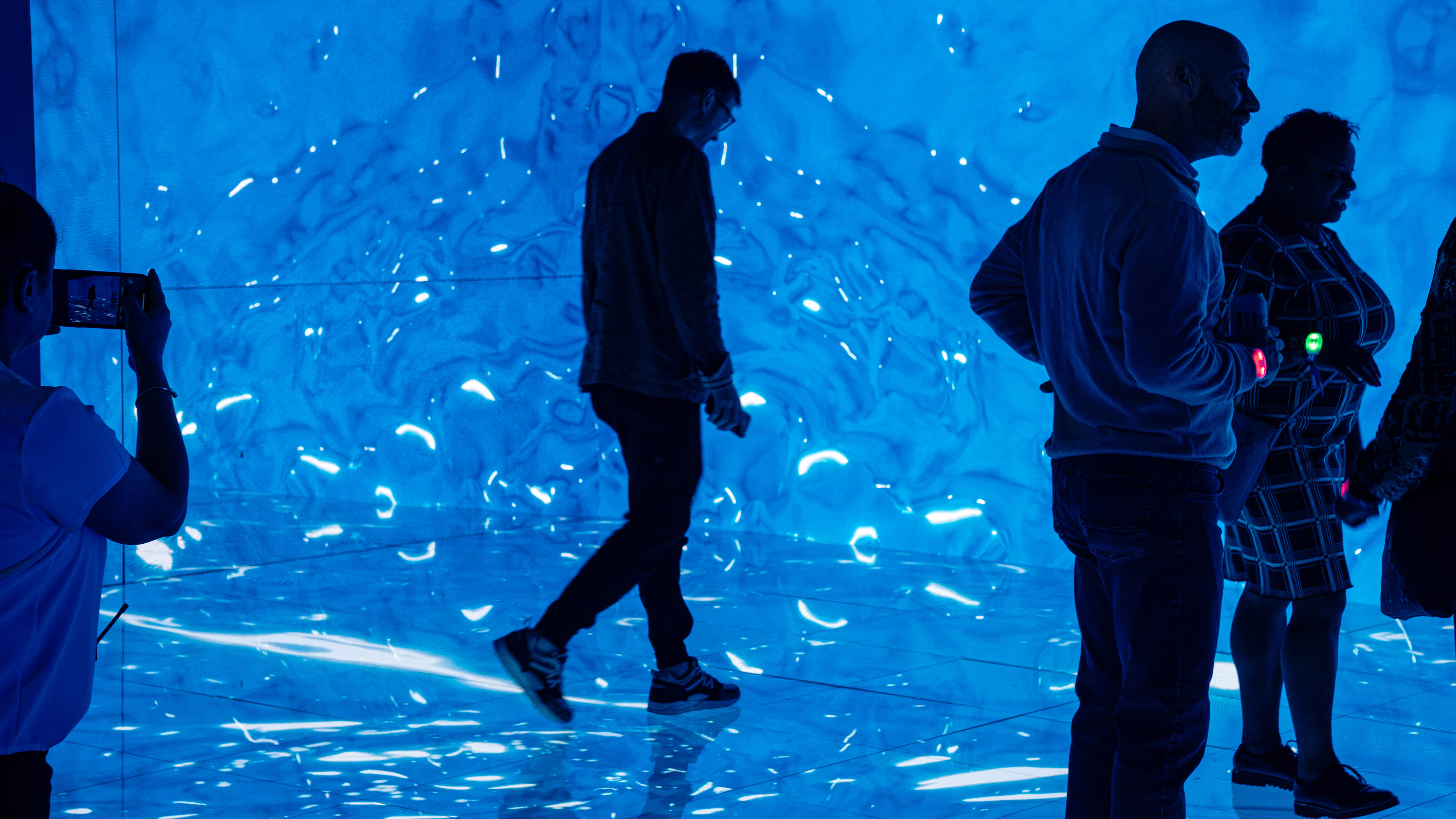 A man walking on an led floor with dynamic water graphics of waves all around him. Other people are also in standing in the room and taking photographys