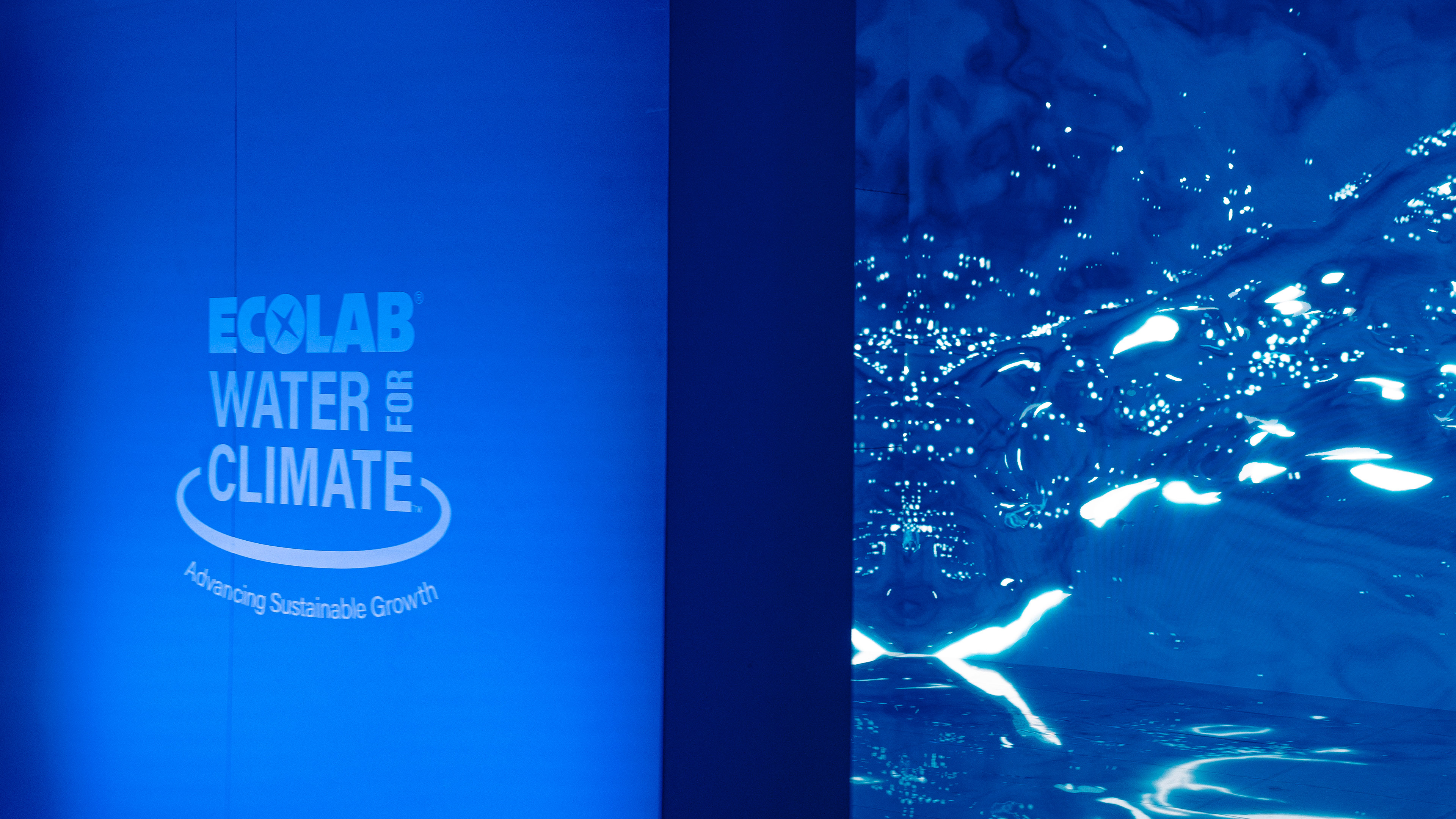 A photo of the entrance to the Ecolab Water for Climate installation at the 100 year anniversary of Ecolab. A blue wall with leds screens filled with simulated water graphics beyond.