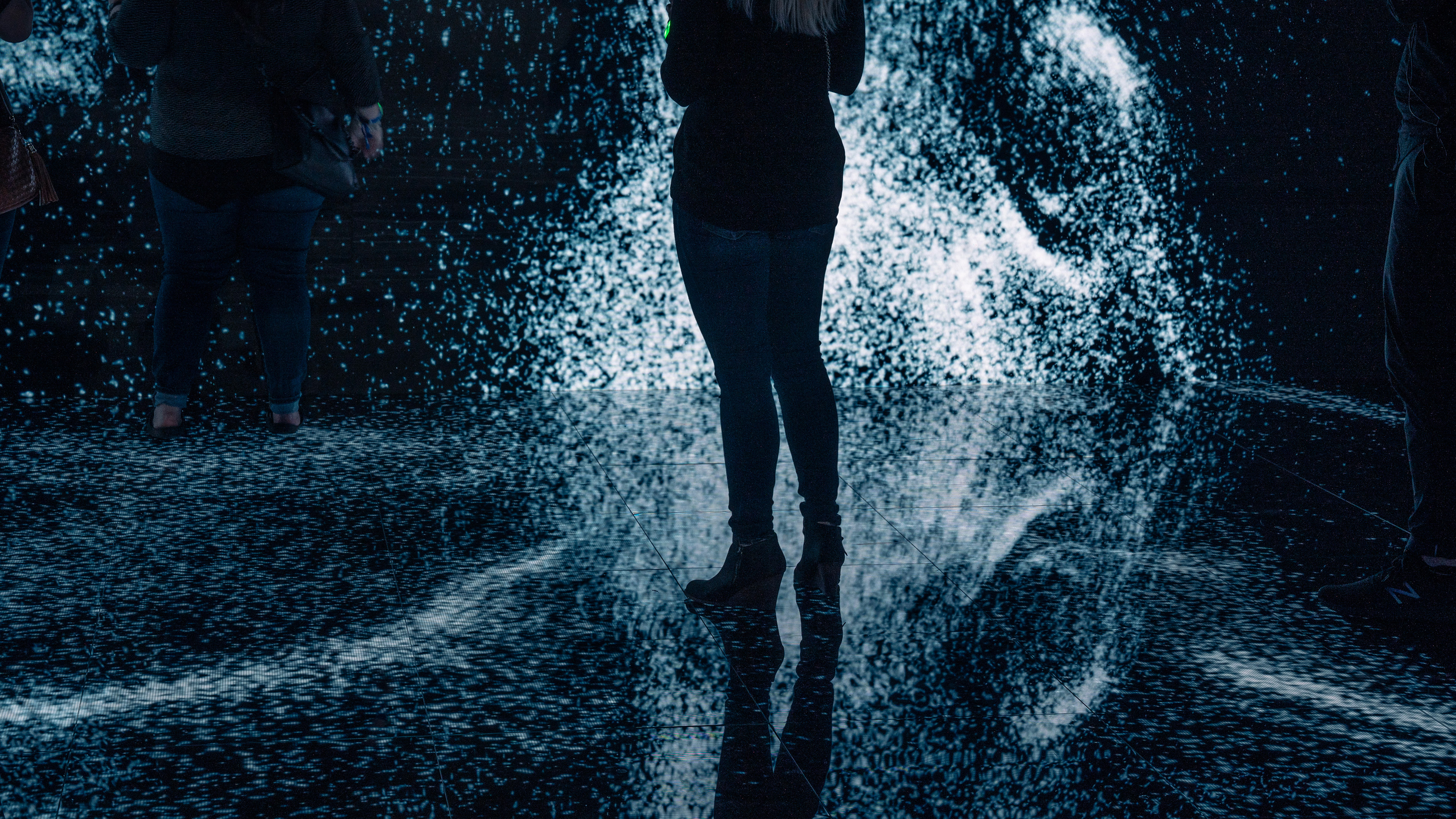 Two people standing in an interactive installation with an led floor and walls. Both are surrounded by generative steam particles