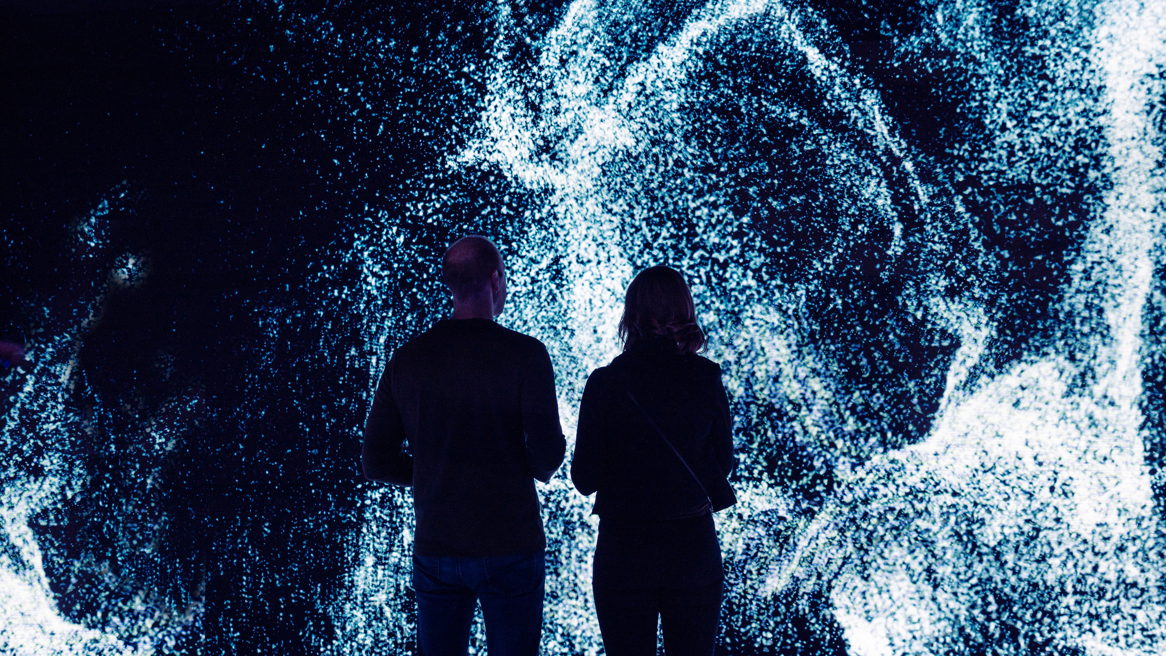 Two people standing in front of a large led screen filled with generative steam particles
