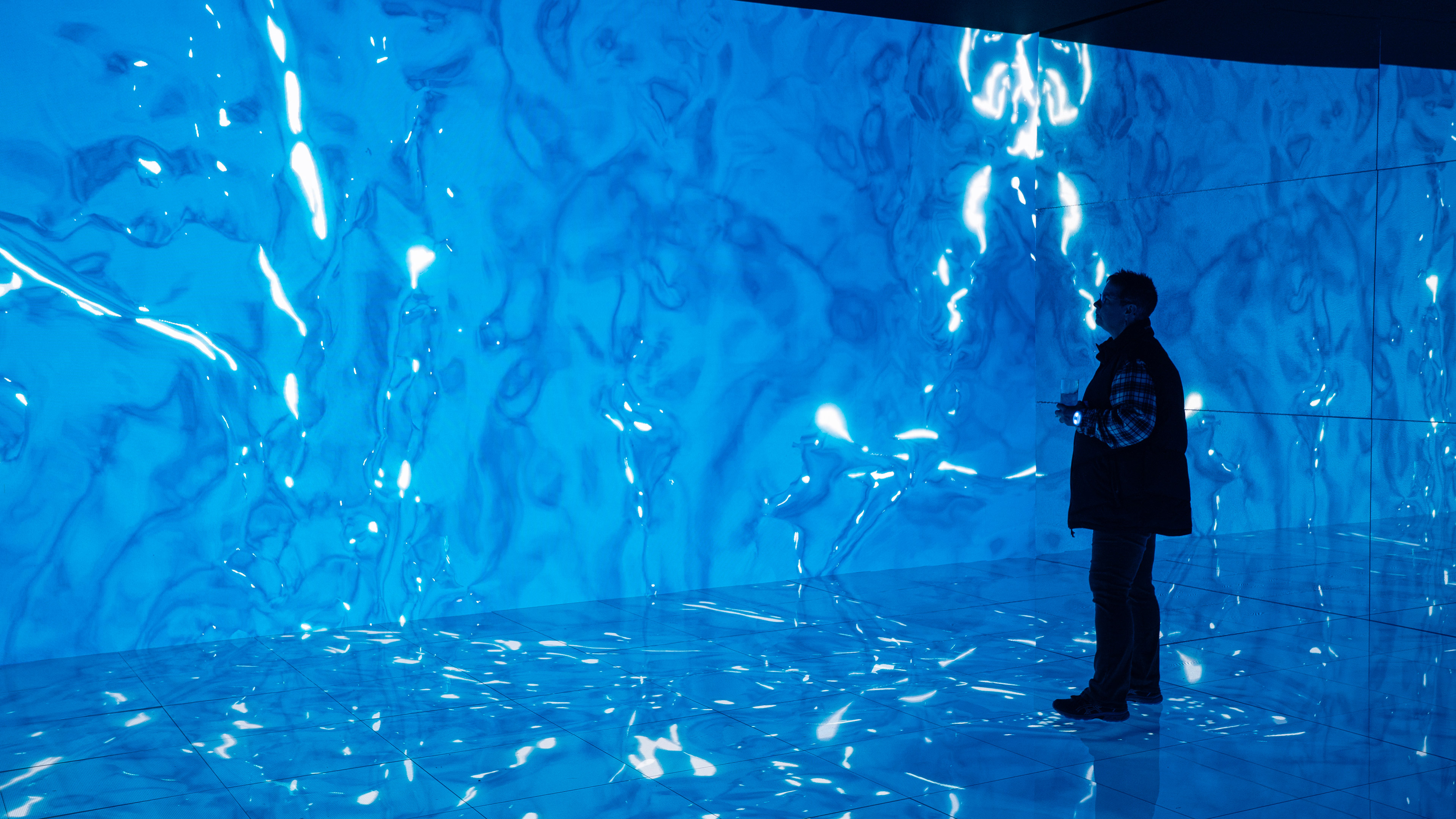 A man standing in an interactive installation with a large led wall and floor and mirrors. The screens are showing an immersive water fluid simulation