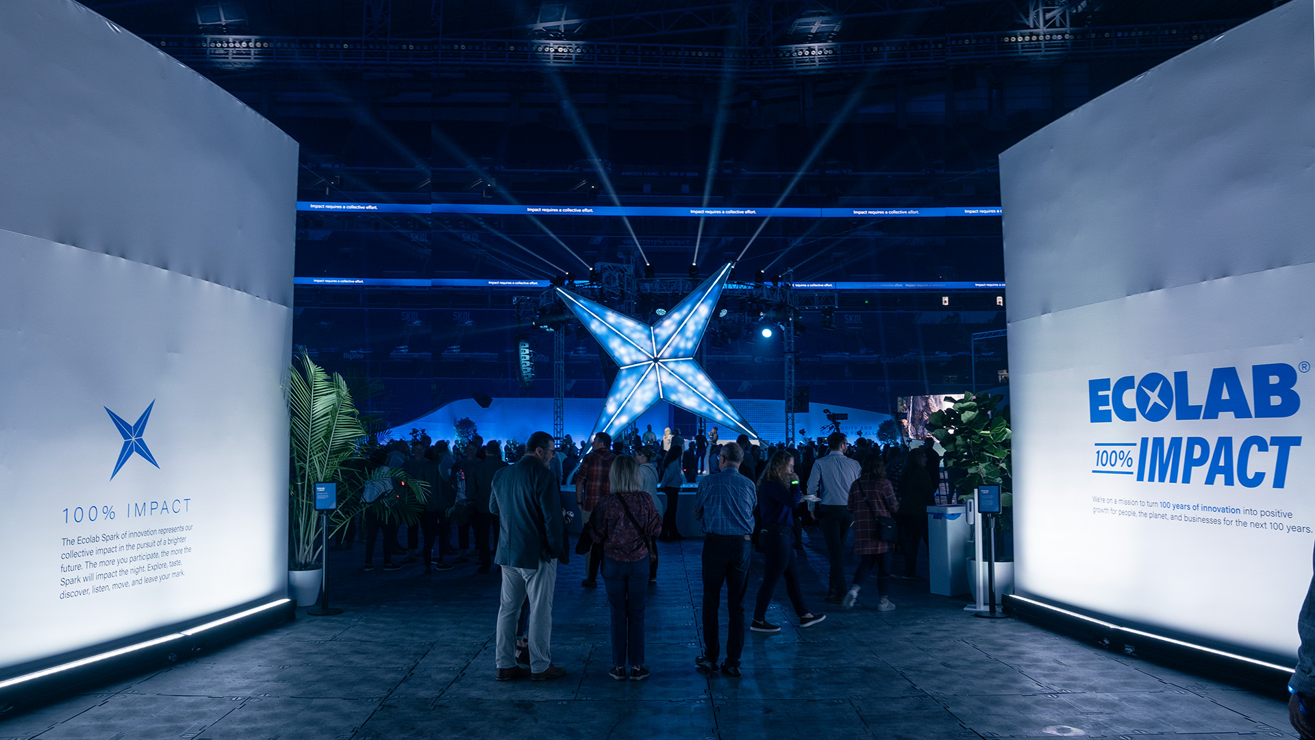 A photo captures the entrance during Ecolab's 100th anniversary, featuring 'The Spark' digital sculpture at its heart. Surrounding it, attendees form a semicircle, with some admiring the sculpture while others engage in conversation, smiles brightening the scene.