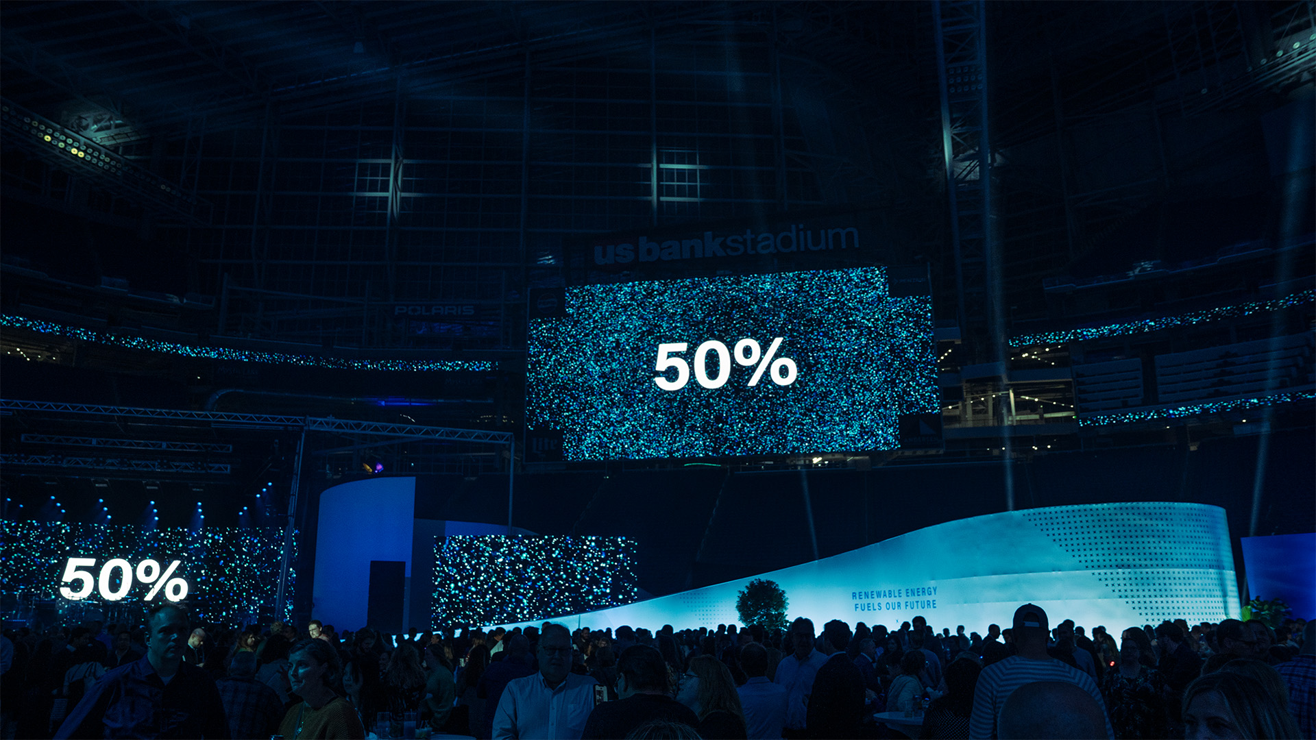 A wide-angle shot of people talking in the stadium. The stage screen at the back is showing "50%" in big font at the center, with a particle background. Another big screen on the back, hanging at the mid air is showing a similar image of percentage